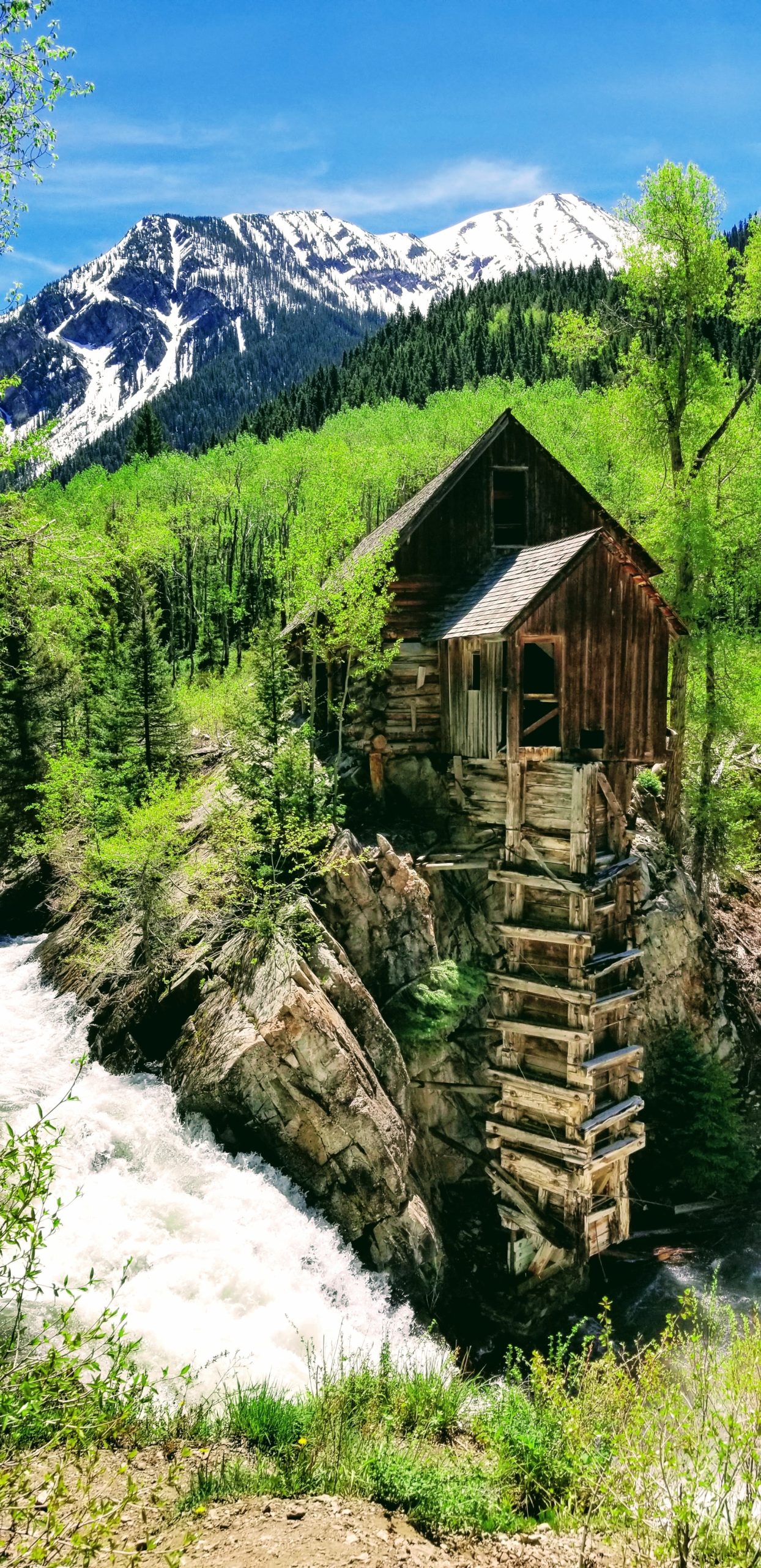 An old, dilapidated wooden mill teetering on a cliff with whitewater rushing below it, surrounded by yellow and green aspens