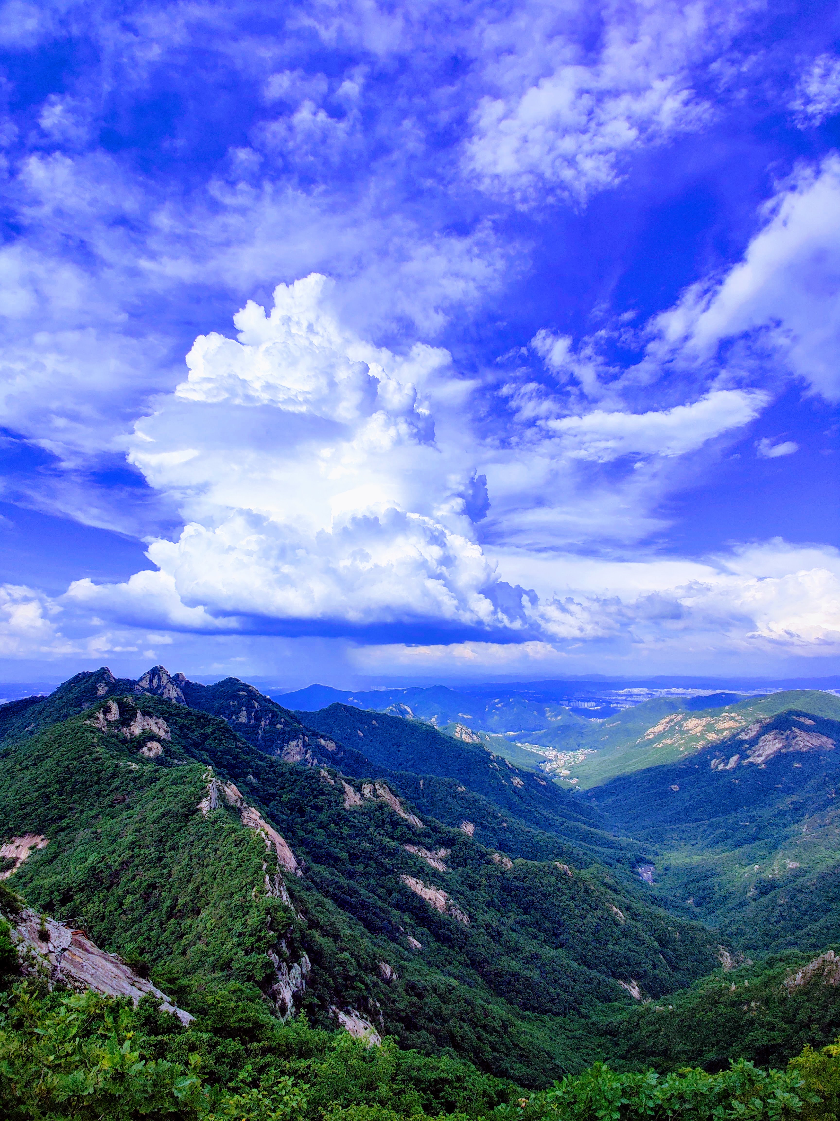a rugged mountainous ridgeline made of tan rock struggles to appear through lush trees and foliage next to a valley as a massive thunderhead is building far in the distance over the mountains of South Korea