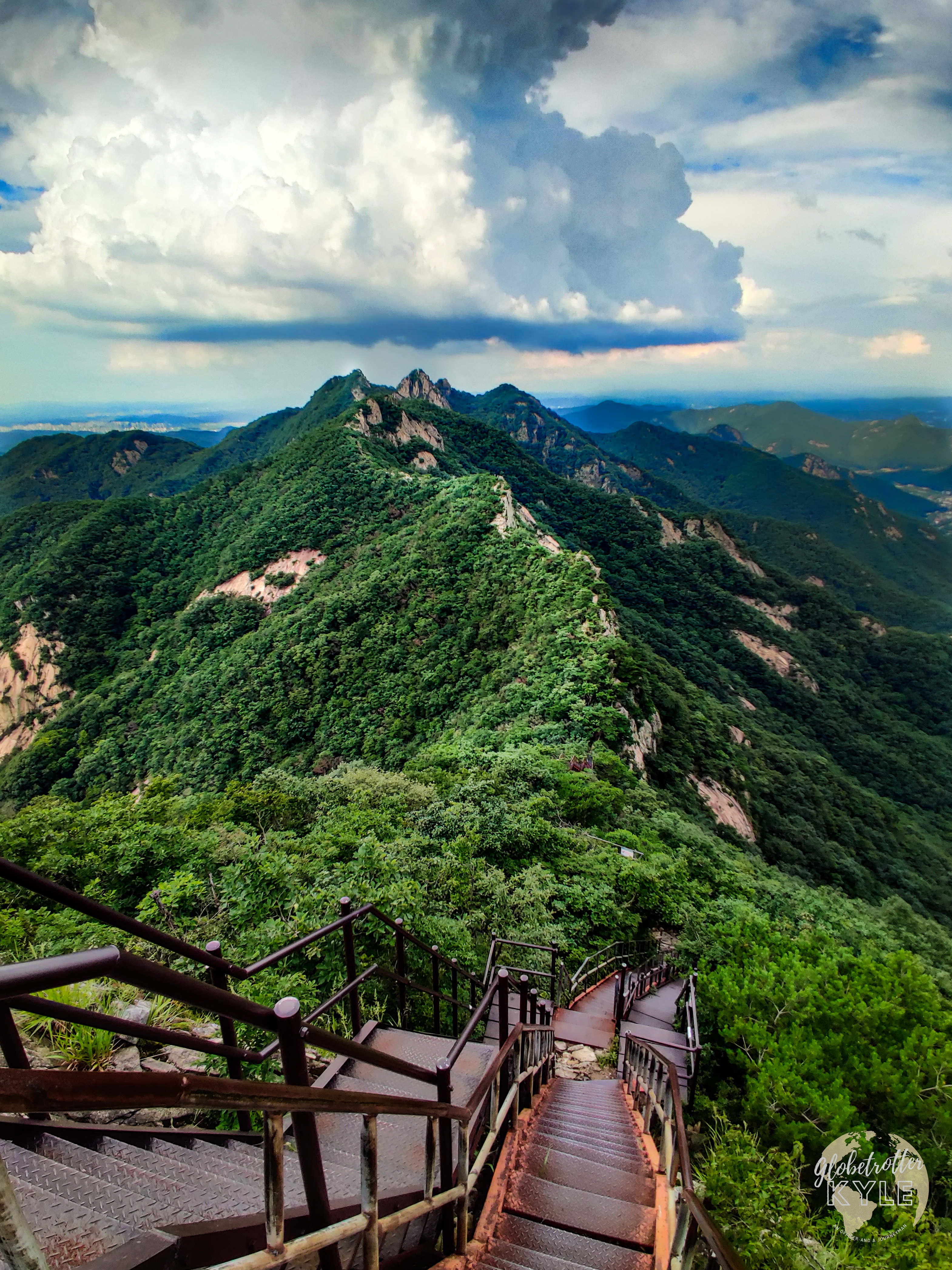 a rugged mountainous ridgeline made of tan rock struggles to appear through lush trees and foliage next to a valley as a massive thunderhead is building far in the distance over the mountains of South Korea