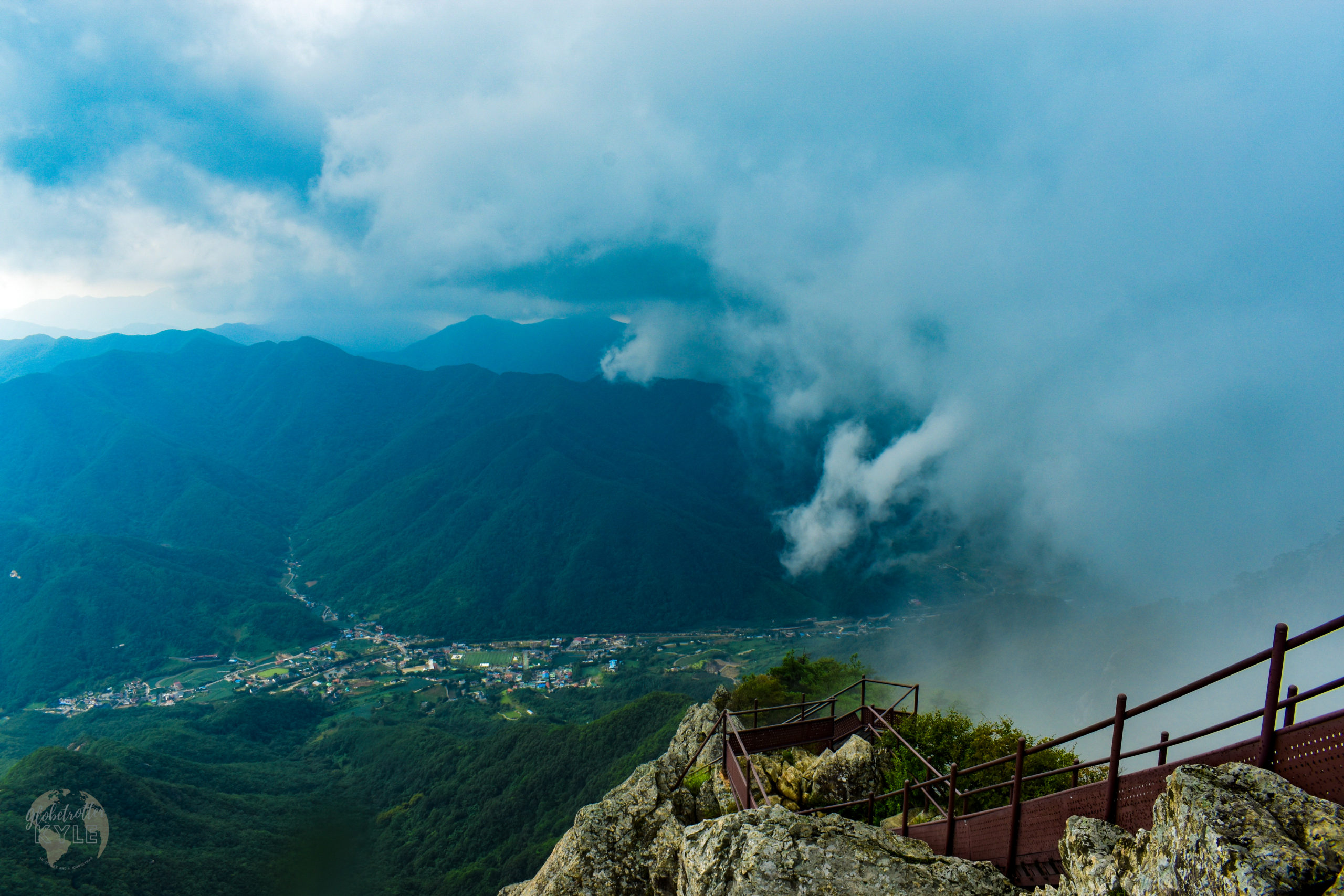 standing atop a mountain in south korea, clouds swarm in from the right as they begin to swallow the viewer's 10 mile visibility range with a mountain range in the distance
