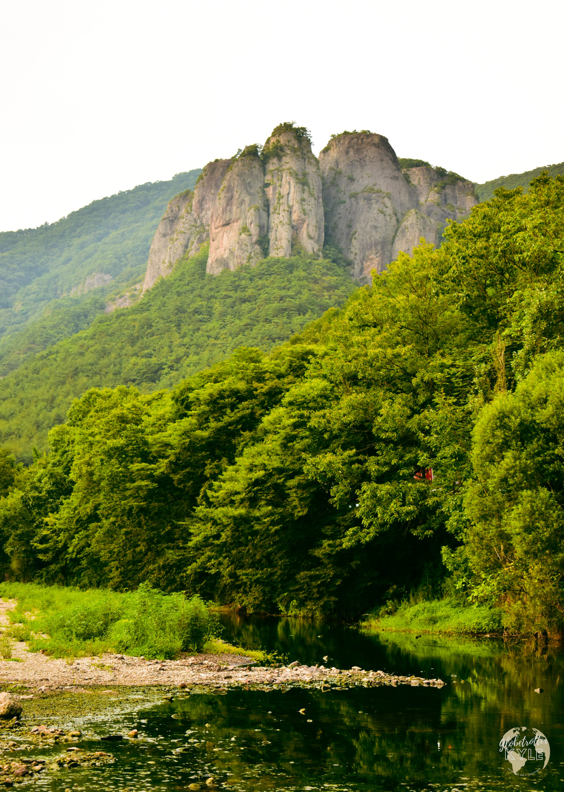 rocks rise above a forested valley in front of a river in south korea juwangsan national park