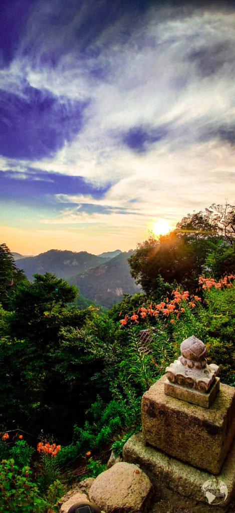 a buddhist temple (Sanggoam) stone column with a lotus leaf carving reflects light from the sun as it sets in the background over a mountain range