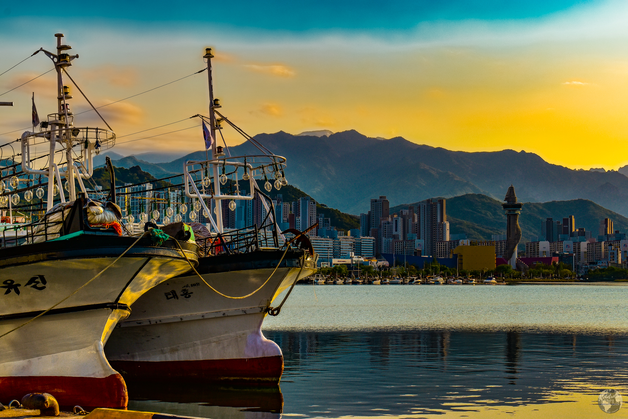 Two boats at rest in the Cheongcho Lake harbor in front of a cityscape below a yellow and orange sky made from the sun setting behind a mountain range