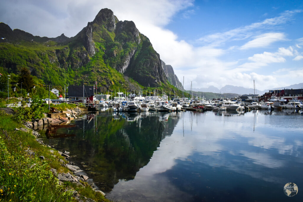 Boats in a marina below a mountain with still water reflectin the image seen when Hiking Lofoten Islands