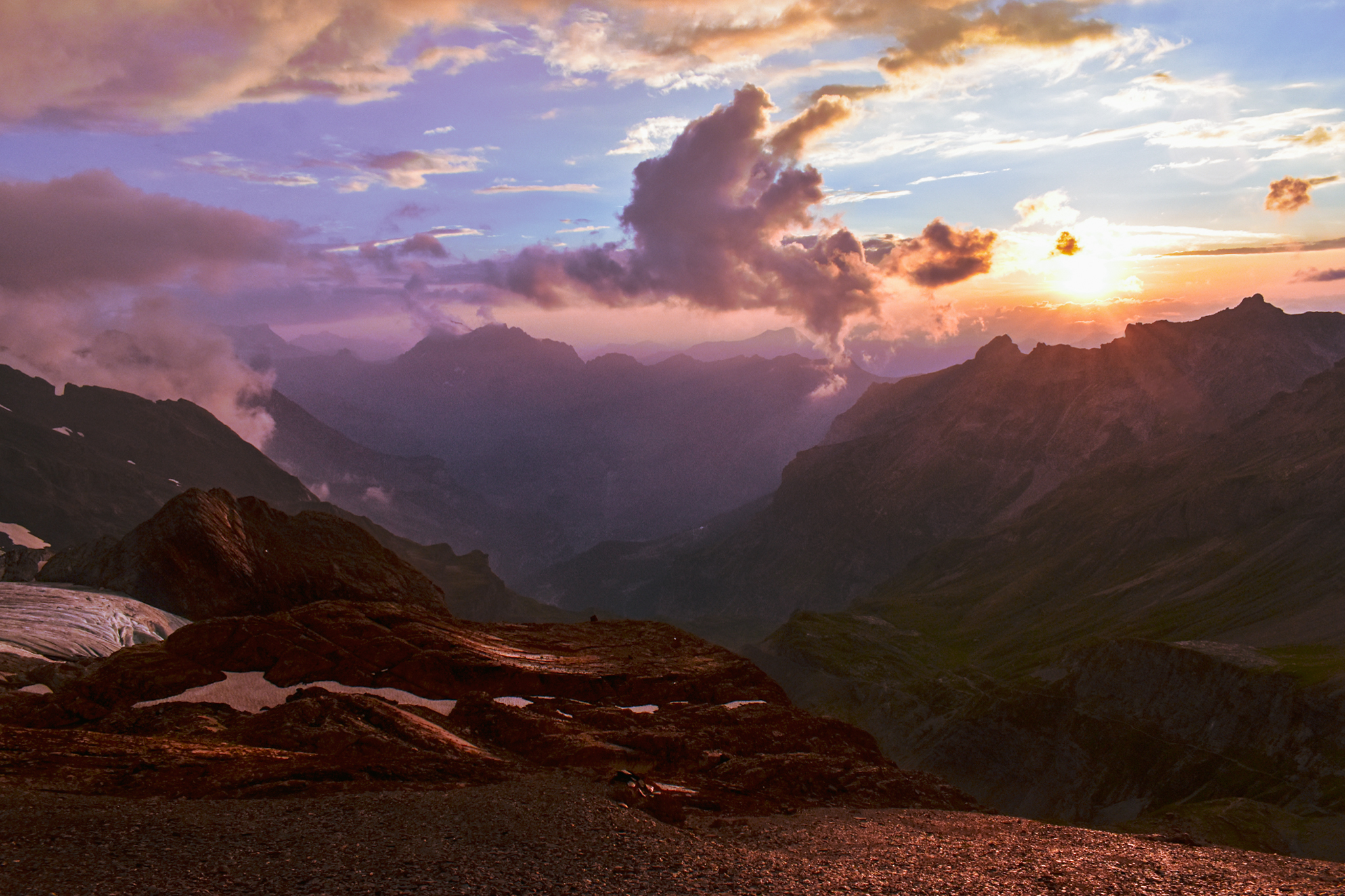 A sunset in the Swiss Alps shows beautiful oranges and yellows in the sky while the mountains in the background appear purple