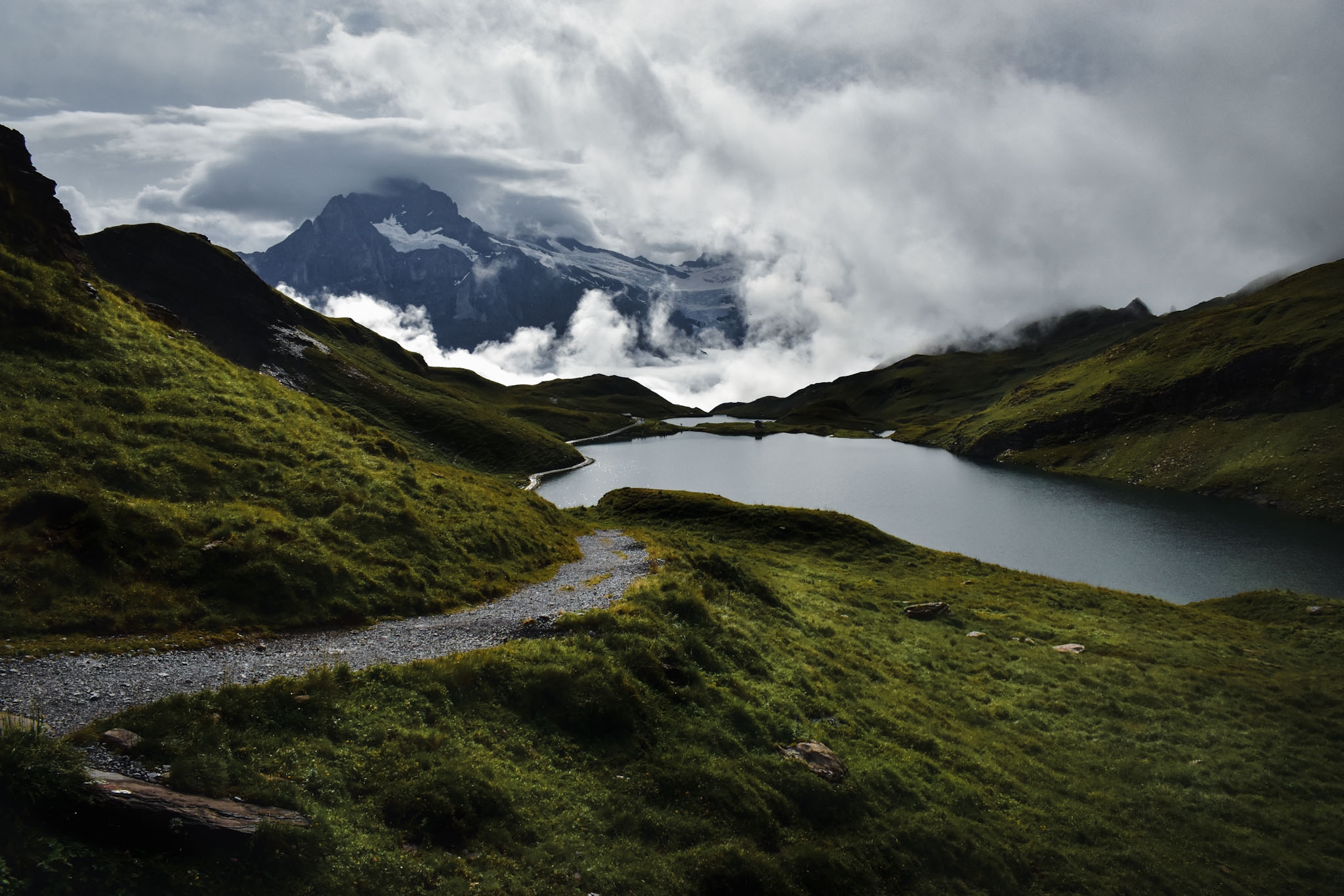 A high alpine lake (Bachalpsee near Berghotel Faulhorn) in the swiss alps with a trail leading to it as a mountain peeks through a clearing in dense clouds while on a hut to hut hike in Switzerland