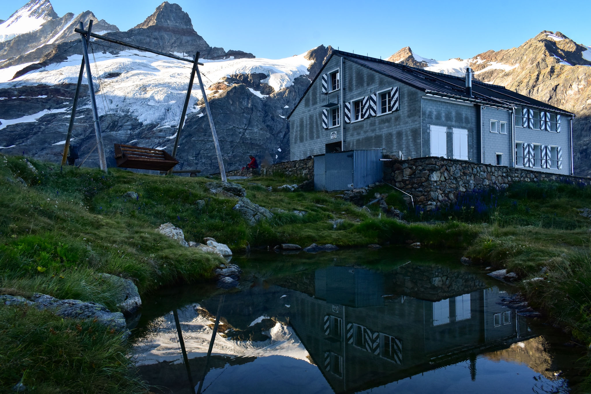 Glecksteinhütte in the Swiss Alps reflects in a high alpine pond with a bench swing next to it, set in front of a white glacier on the side of a mountain as seen while doing a hut to hut hike in Switzerland