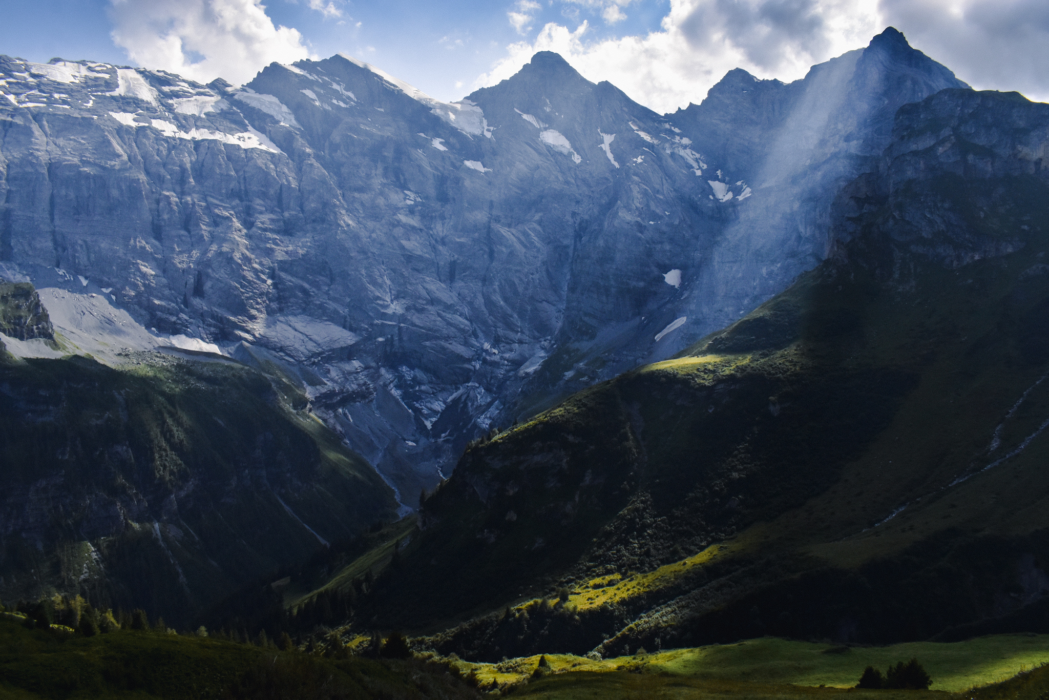 a ray of sun shines through the clouds and highlights a green pasture in front of a large mountain range in Switzerland