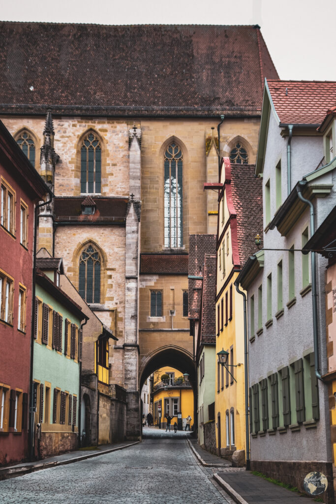 A cobblestone street going under a church archway with buildings and people walking in Rothenburg ob der Tauber
